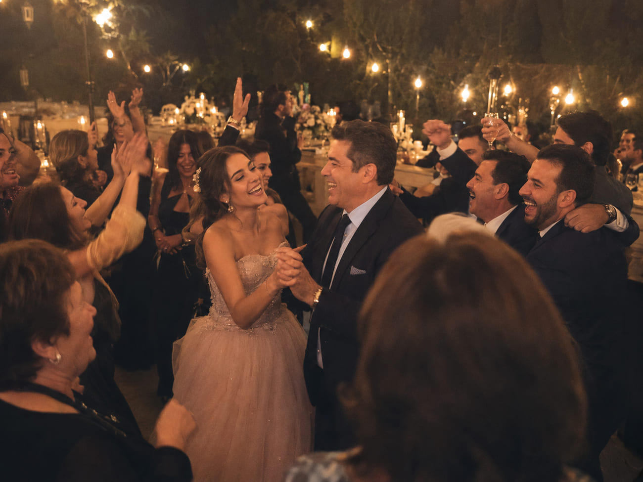 Pista de baile llena: la quinceañera baila riendo con su papá mientras la familia canta, aplaude y brinda bajo faroles cálidos