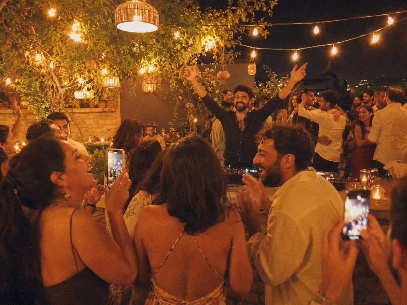 DJ riendo con los brazos en alto mientras los invitados cantan y otros bailan felices al fondo entre luces cálidas