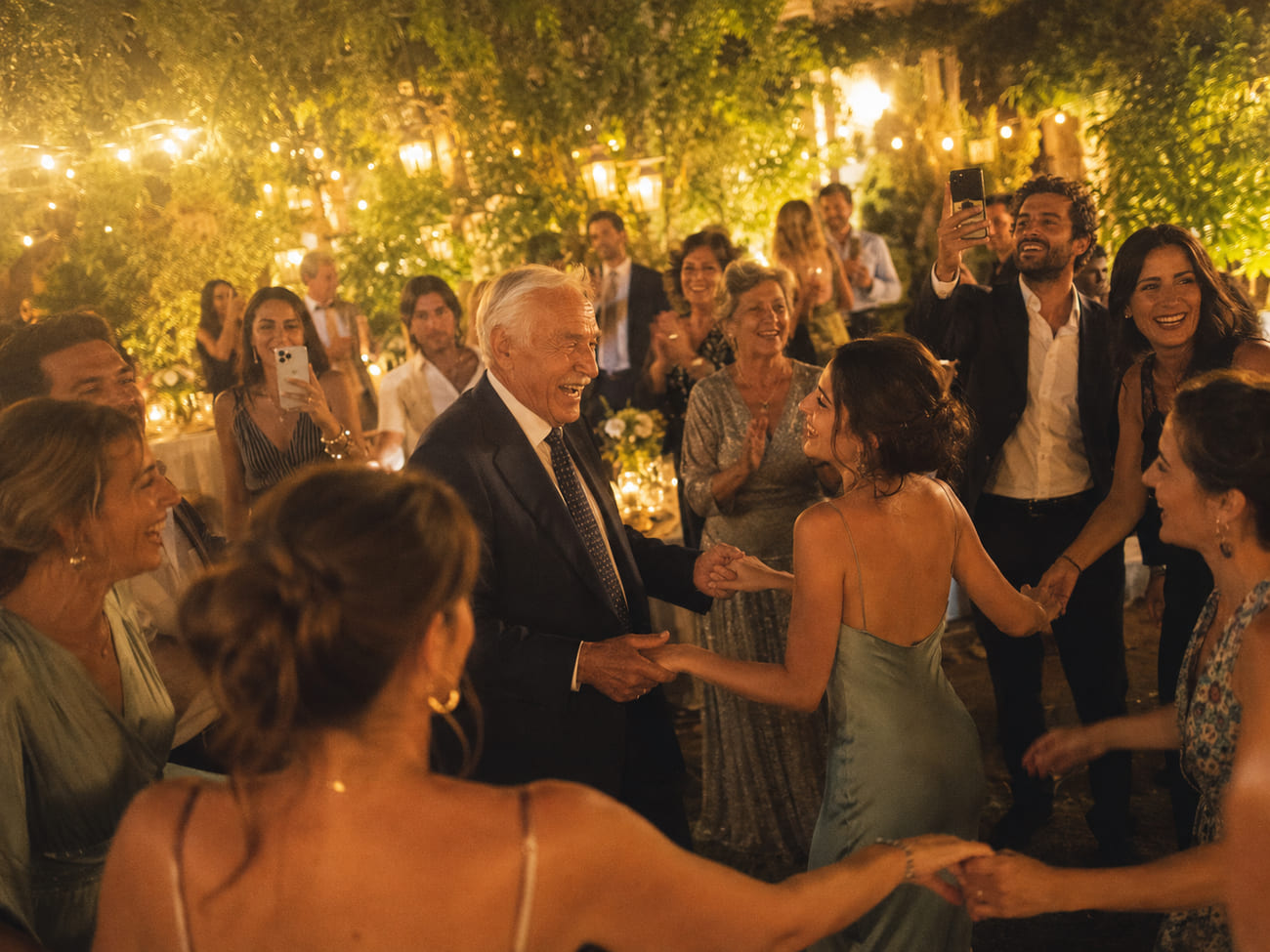 Círculo de baile con abuelos y jóvenes riendo tomados de las manos, invitados brindando al fondo entre faroles dorados