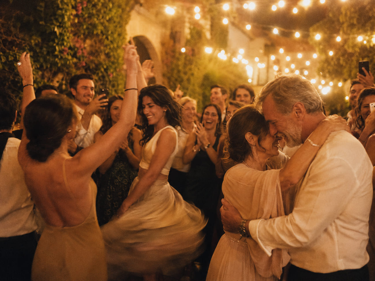 Invitados elegantes bailando radiantes con los brazos en alto en un patio bajo guirnaldas cálidas, con una pareja mayor abrazada y todos cantando y riendo al fondo.