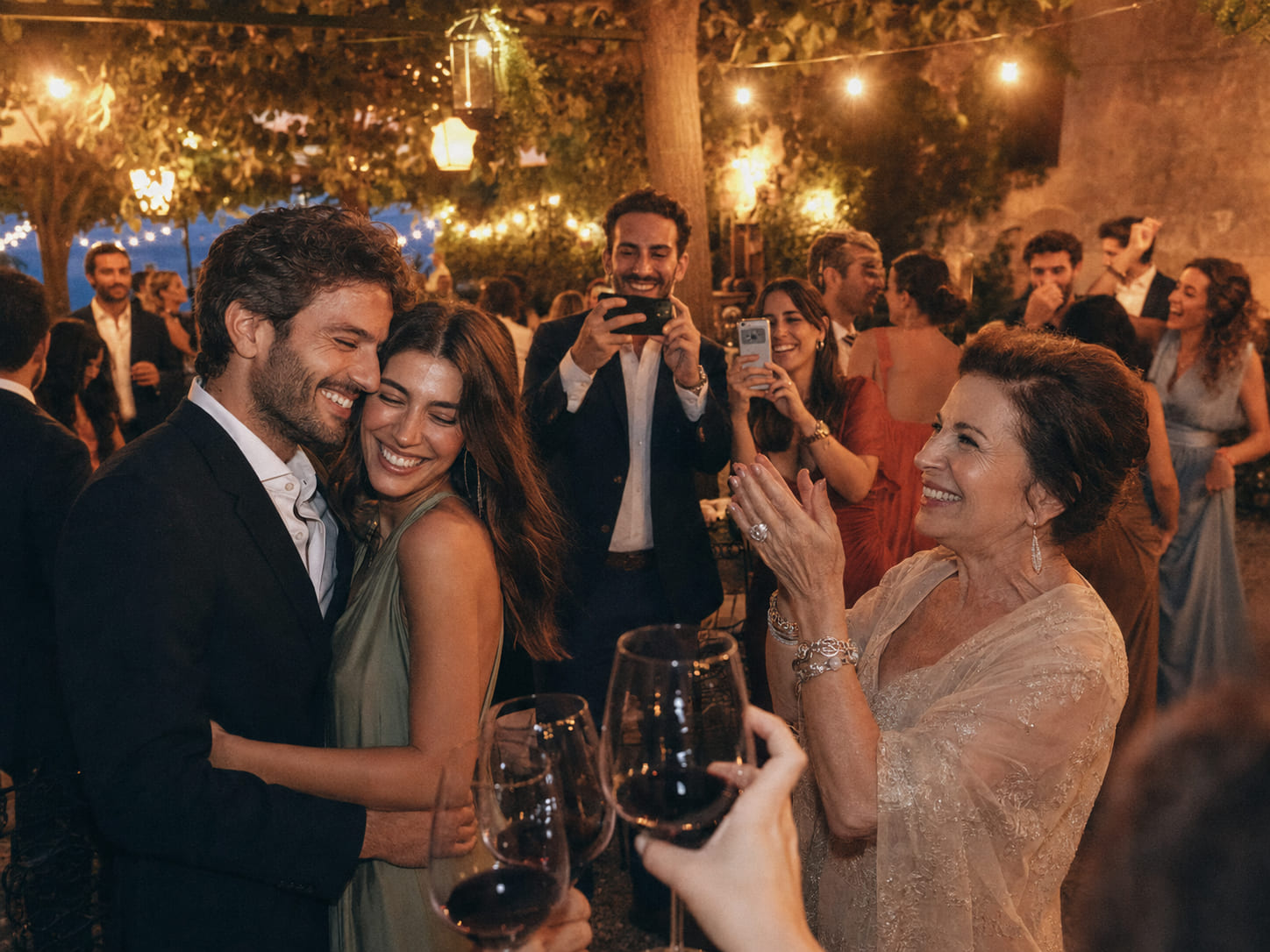 Invitados elegantes riendo y brindando con copas llenas en un jardín con guirnaldas al atardecer, mientras al fondo todos bailan y ríen entre faroles desenfocados.