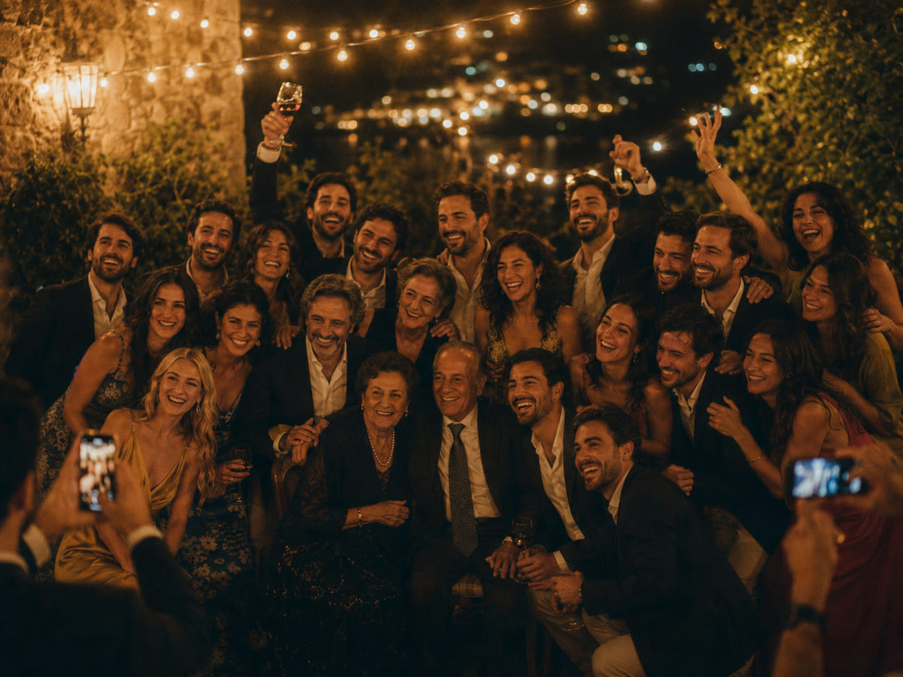 Foto grupal de cierre en un patio con guirnaldas: veinte invitados abrazados en dos filas, riendo con los brazos en alto y abuelos encantados al centro.