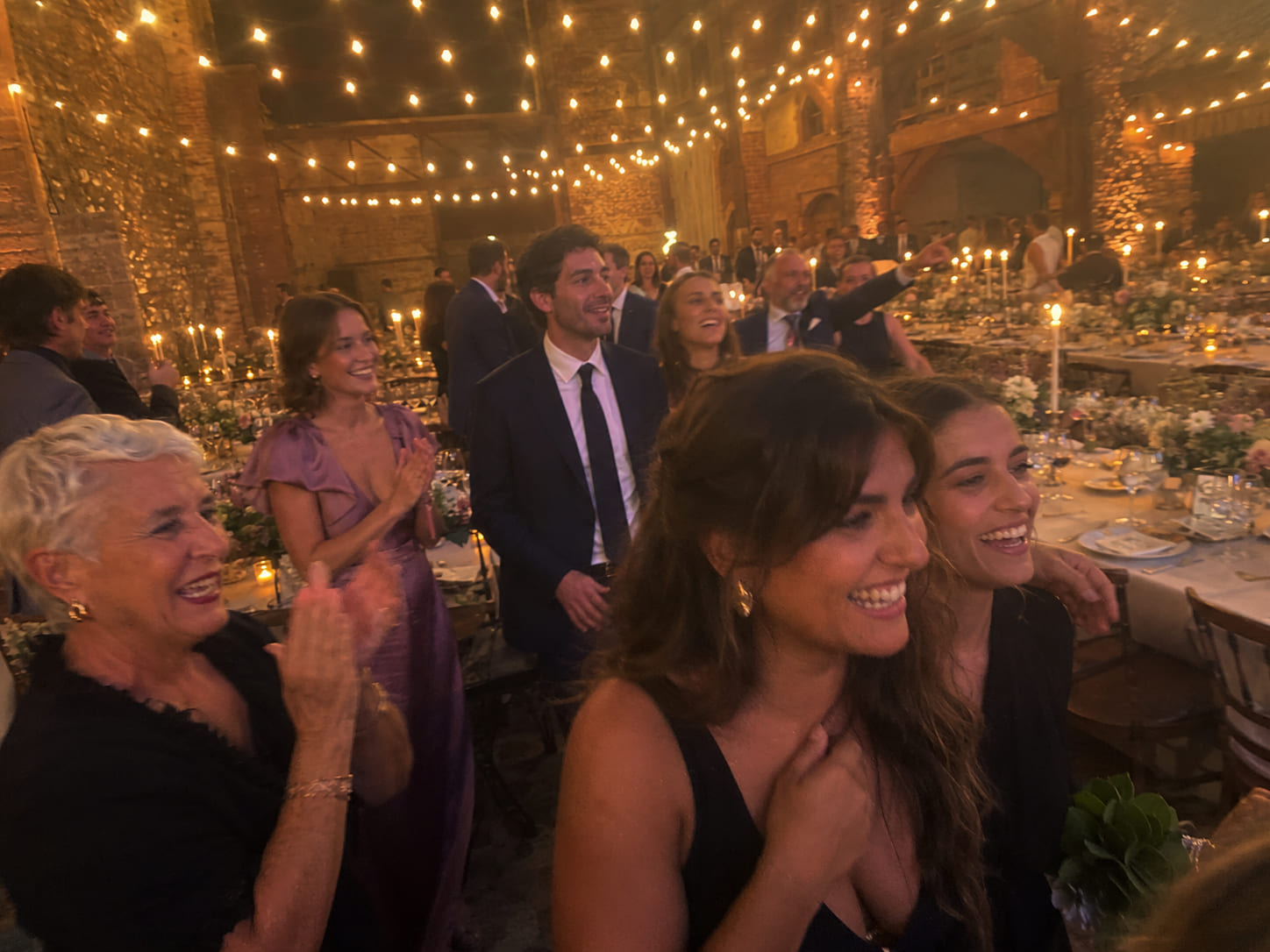 Invitados elegantes entran riendo y aplaudiendo a un salón con velas, flores y mesas servidas