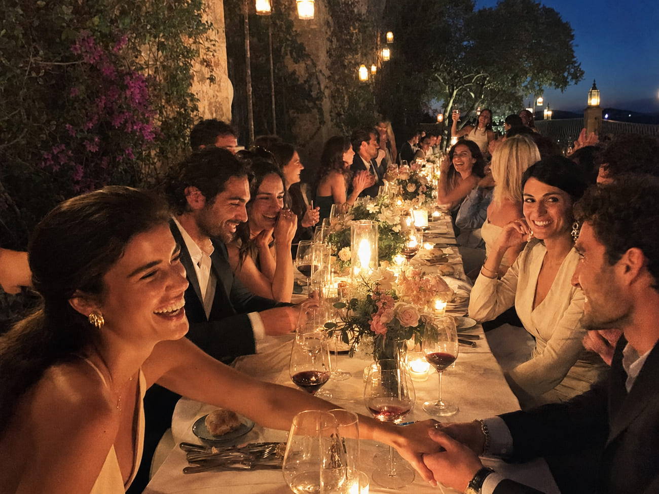 Mesa larga con velas, flores frescas y copas llenas, invitados elegantes riendo juntos mientras otros brindan al fondo.