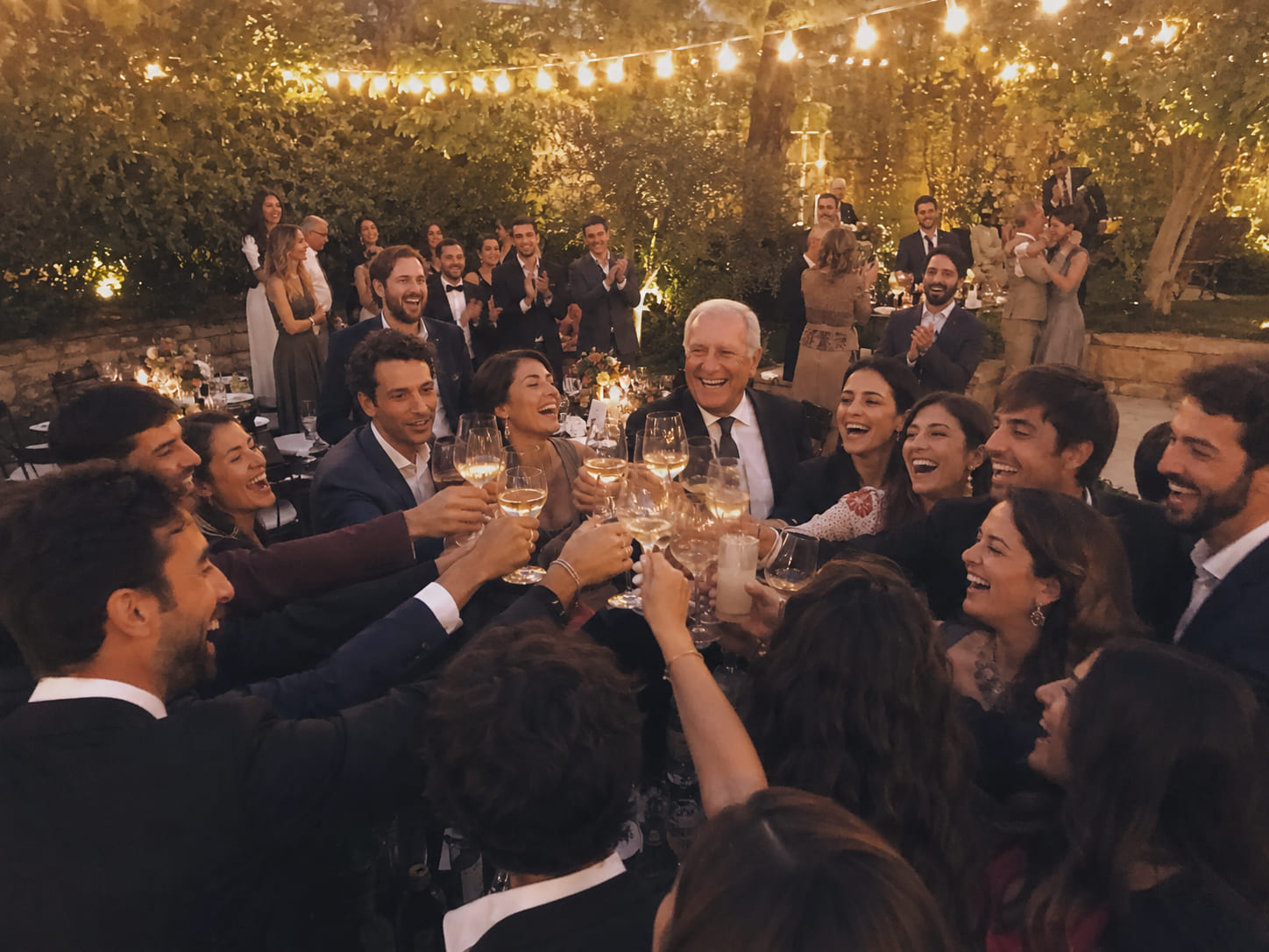 Invitados elegantes brindan riendo en un jardín al atardecer, con guirnaldas encendidas y todos celebrando al fondo.
