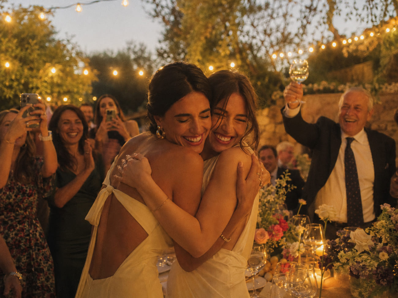 Dos invitadas se abrazan felices en un jardín con guirnaldas encendidas mientras amigas aplauden riendo y un señor mayor elegante celebra con su copa en alto