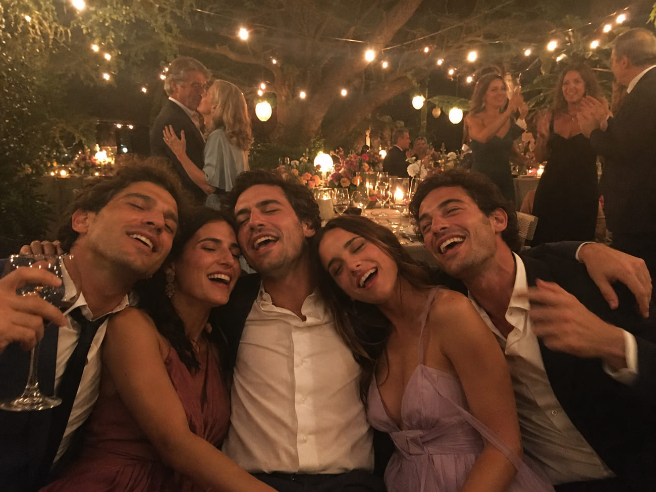 Cinco invitados elegantes sentados juntos en un banco bajo guirnaldas encendidas, riendo y cantando abrazados, mientras una pareja baila y otros brindan al fondo